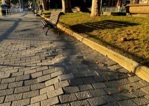 A park bench positioned on a sidewalk with an uneven surface nearby.
