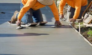 Two men in orange shirts repair a concrete sidewalk, focused on their work on a sunny day.