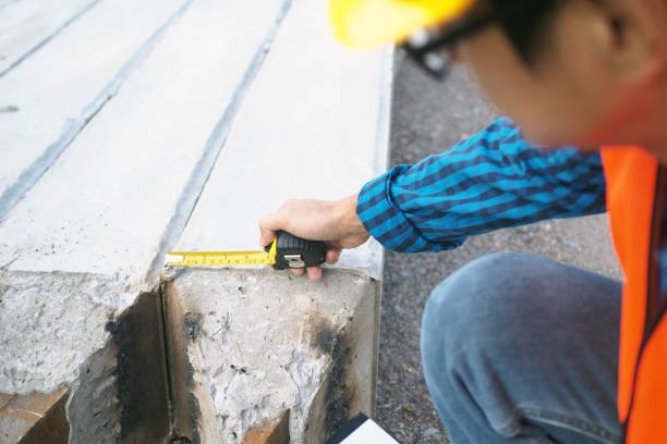 A construction worker in a hard hat and safety vest uses a tape measure on a piece of wood for walkway repairs.
